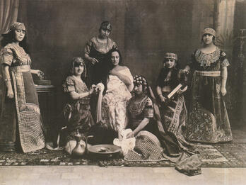 Photograph of several women in dresses and headdresses with rug and cooking implements on the floor.