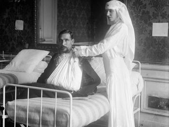 Photograph of woman in veil standing next to bed where man sits with arm in sling.