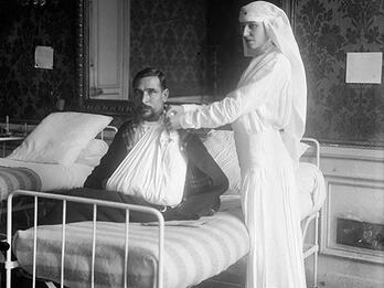 Photograph of woman in veil standing next to bed where man sits with arm in sling.