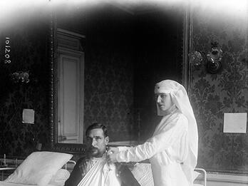 Photograph of woman in veil standing next to bed where man sits with arm in sling.