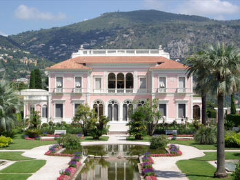 Photograph of house surrounded by gardens and fountain and flanked by two tall palm trees, with mountains in background.