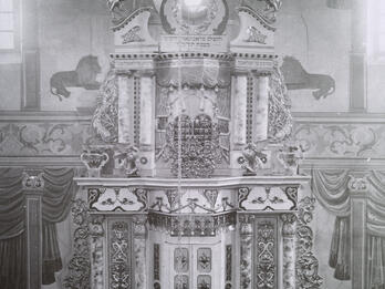 Photograph of large, ornate structure decorated with flourishes, lions, and vines, with three men in hats and beards standing in front of two closed doors at the bottom, and several other figures sitting in benches in foreground.