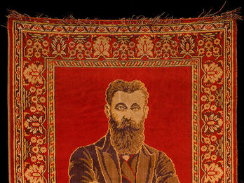 Herzl Rug Rug depicting a bearded man with crossed arms on top half, and two columns and hanging lamp on bottom half, surrounded by decorative border.