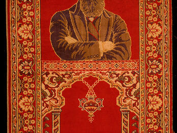 Rug depicting a bearded man with crossed arms on top half, and two columns and hanging lamp on bottom half, surrounded by decorative border.