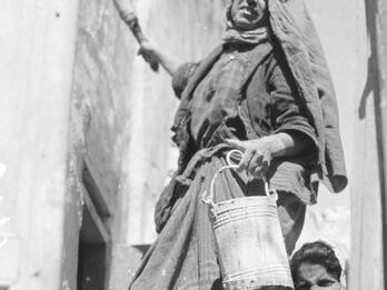 Photograph of woman in headscarf looking toward camera, holding bucket and paintbrush next to wall, with woman behind her looking at camera.