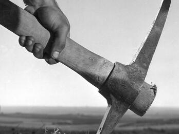 Photograph of a hand holding a garden pick with field in background.