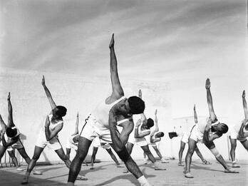 Photograph of rows of men in athletic clothing bending at the waist with their right arms pointed up and their left arms bent in an angle toward their face.