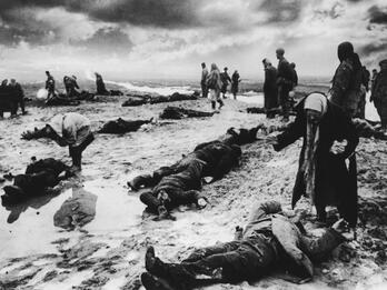 Photograph of people standing in a field upon which lie dead bodies.