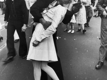 Photograph of sailor grabbing and kissing a woman in a busy city street with people and buildings in background. 