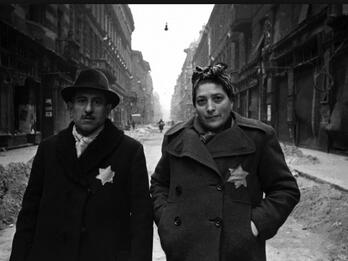 Budapest Ghetto Photograph of man and woman standing in the middle of a city street wearing jackets with yellow Stars of David on breast.