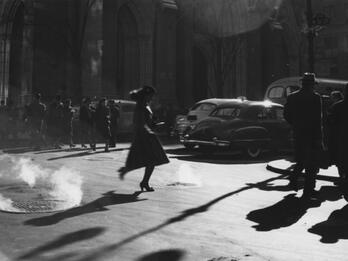 Photograph of woman in a dress and hat walking across a city street with cars, people, and buildings in the background.