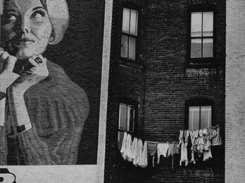 Photograph depicting painted advertisement of a woman on wall of brick building next to a clothesline on adjacent wall.