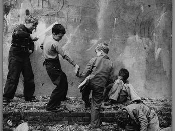 Photograph of five young boys standing and sitting in various poses on crumbling foundation of a wall with swastika drawn on it. One boy points a flat wooden stick at another boy, who is giving a metal object to a seated boy. 