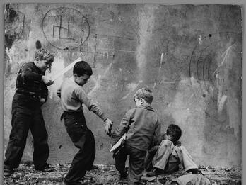 Photograph of five young boys standing and sitting in various poses on crumbling foundation of a wall with swastika drawn on it. One boy points a flat wooden stick at another boy, who is giving a metal object to a seated boy. 