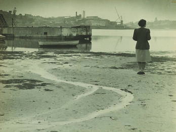 Photograph of a woman standing on a wide beach and facing away from the viewer toward a river.