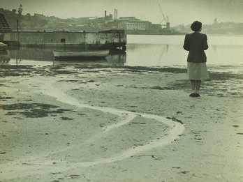 Photograph of a woman standing on a wide beach and facing away from the viewer toward a river.