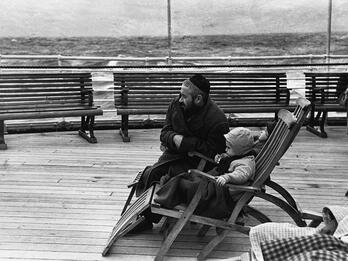 Coming to America Photograph of man in a skullcap kneeling next to a child sitting in a wooden chair on boat deck, gazing into the distance to the left of the viewer.