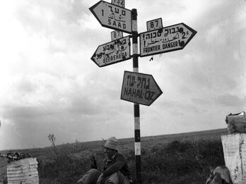 Outdoor photograph of signpost with signs pointing different directions with town names and information in Hebrew, English, and Arabic, and smiling man holding a rifle sitting on the ground beneath the sign.