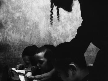 Hebrew Lesson, Brooklyn, New York Photograph of man in a hat and sidelocks bending over a desk where three boys are reading Hebrew.