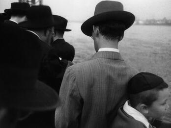 Photograph of men in hats and suits and one boy in skullcap outside with their backs turned away from the camera, looking toward the river.