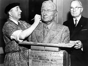 Photograph of man in beret and decorated short-sleeved jacket using a tool on a clay model bust of a man in suit and wearing glasses, who is standing behind the bust. 