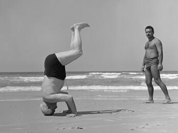 Photograph of two shirtless men on the beach, one doing a headstand with legs bent facing to the right of the viewer and the other man facing the viewer on the right side of the photograph.