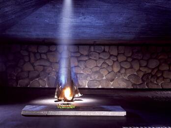 Memorial depicting flame burning from a base and illuminating a wall with an angular roof and basalt walls, with place names in English and Hebrew on the floor.