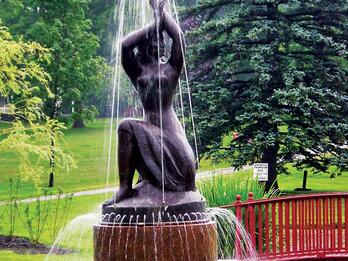 Bronze sculpture and fountain in the shape of a partially nude woman, resting on one knee and raising her arms above her head, set in a park with trees and a bridge in the background. 