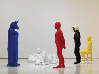 Mixed media sculpture featuring six figures in various poses: a male and a female figure reclining on the ground, a woman seated in a chair to the right, a man with a donkey mask standing to the left, Superman standing in center, and a woman standing in the background.