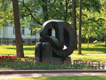 Monument depicting curved, stylized figure with head in hands set among flowers in a grassy park with a house in the background.