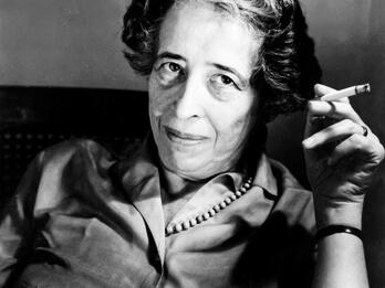 Portrait photograph of a woman looking at the camera and holding up a cigarette in her left hand.