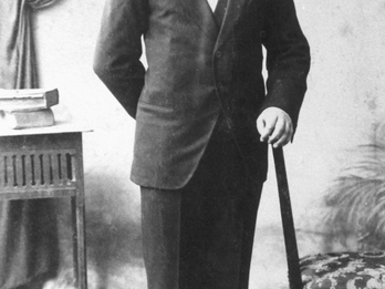 Photograph of man in suit with one arm on a walking stick. 