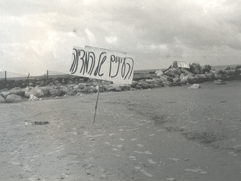 The Eyes of the Nation Photograph of Hebrew sign on empty beach with a rock jetty in background.