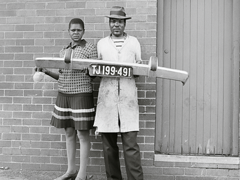 Photograph of man and woman standing and holding a car bumper in front of a brick wall.  