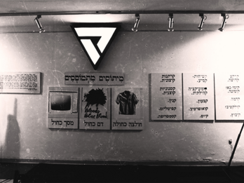 Photograph of wall with a mannequin to the left, posters with Hebrew text and an illuminated triangle on the wall, and a flag to the right.