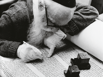 Scribe, N.Y.C. Photograph depicting man in kippah, gloves, and glasses and holding a pen examining a Torah scroll upon which sits two tefillin boxes.
