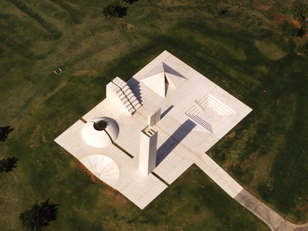 Concrete sculpture featuring a pyramid, water channel, tower with wind flutes, and a dome with olive tree in center set in a field. 
