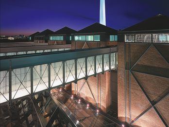 Photograph of brick building exterior with skywalks over a smaller building below with a metal and glass roof and a tall monolith in the background.