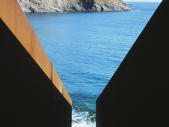Passages, Homage to Walter Benjamin Photograph of metal sculpture in the shape of a V through which the ocean and rocky outcropping beyond is visible.