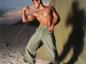 Photograph of man in kippah with shirt off flexing his bicep outside of tent. 