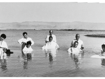 Photograph featuring seven barbers standing in the ocean and cutting hair of seven men floating in front of them. 