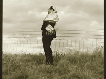 Full body photograph depicting man in kippah and prayer shawl standing in a field next to fence, facing to the side.