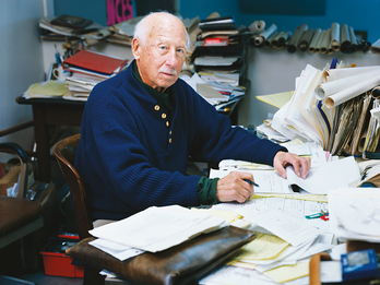 Photograph featuring older man facing viewer and seated at cluttered desk.