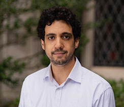 A man with short, curly dark hair looks directly at the camera, an ivy-covered wall visible behind him.