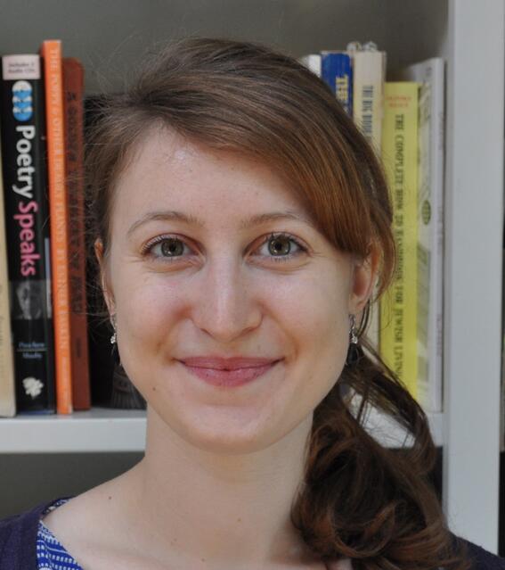 A woman with her hair pulled back looks directly at the camera. Part of a bookshelf is visible behind her.