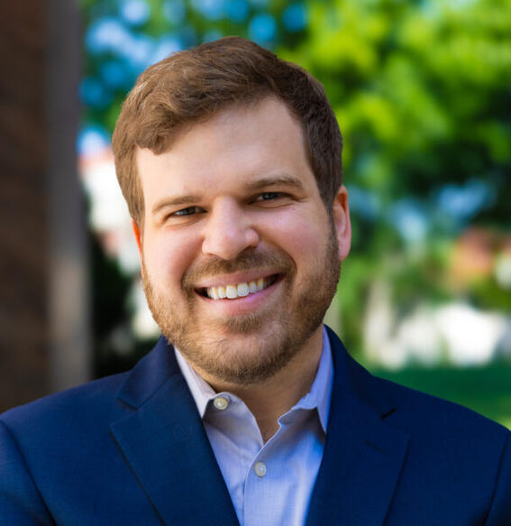A man with short brown hair, beard and mustache, smiles at the camera; he wears a light blue shirt and navy blazer.