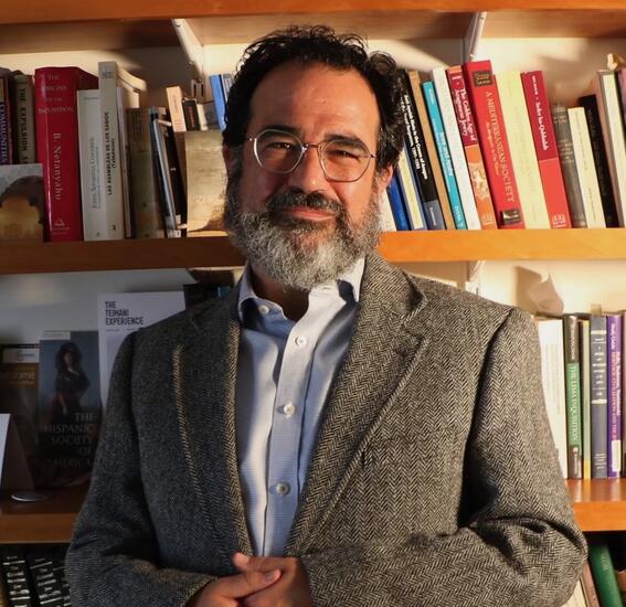 A man with short dark hair, graying beard, glasses, a light blue shirt, and a tweed blazer looks at the camera; a shelf of books is behind him.