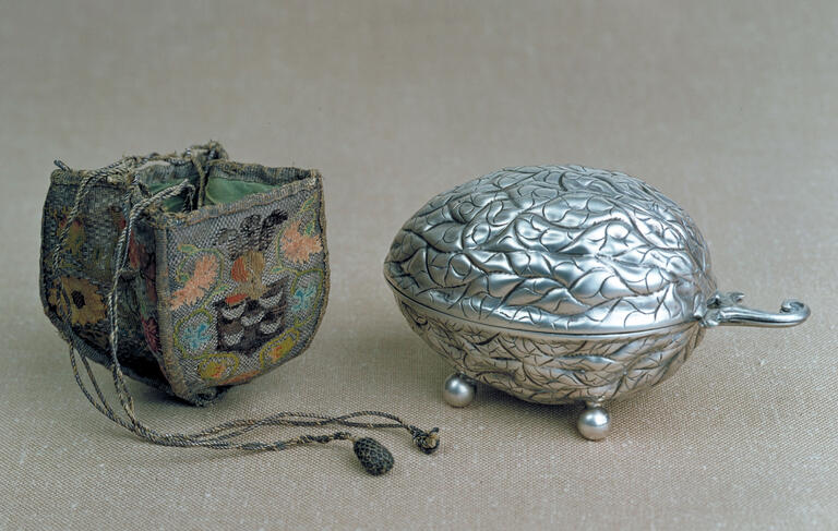 Silver oval container of decorative leaf design and woven, embroidered case next to it.