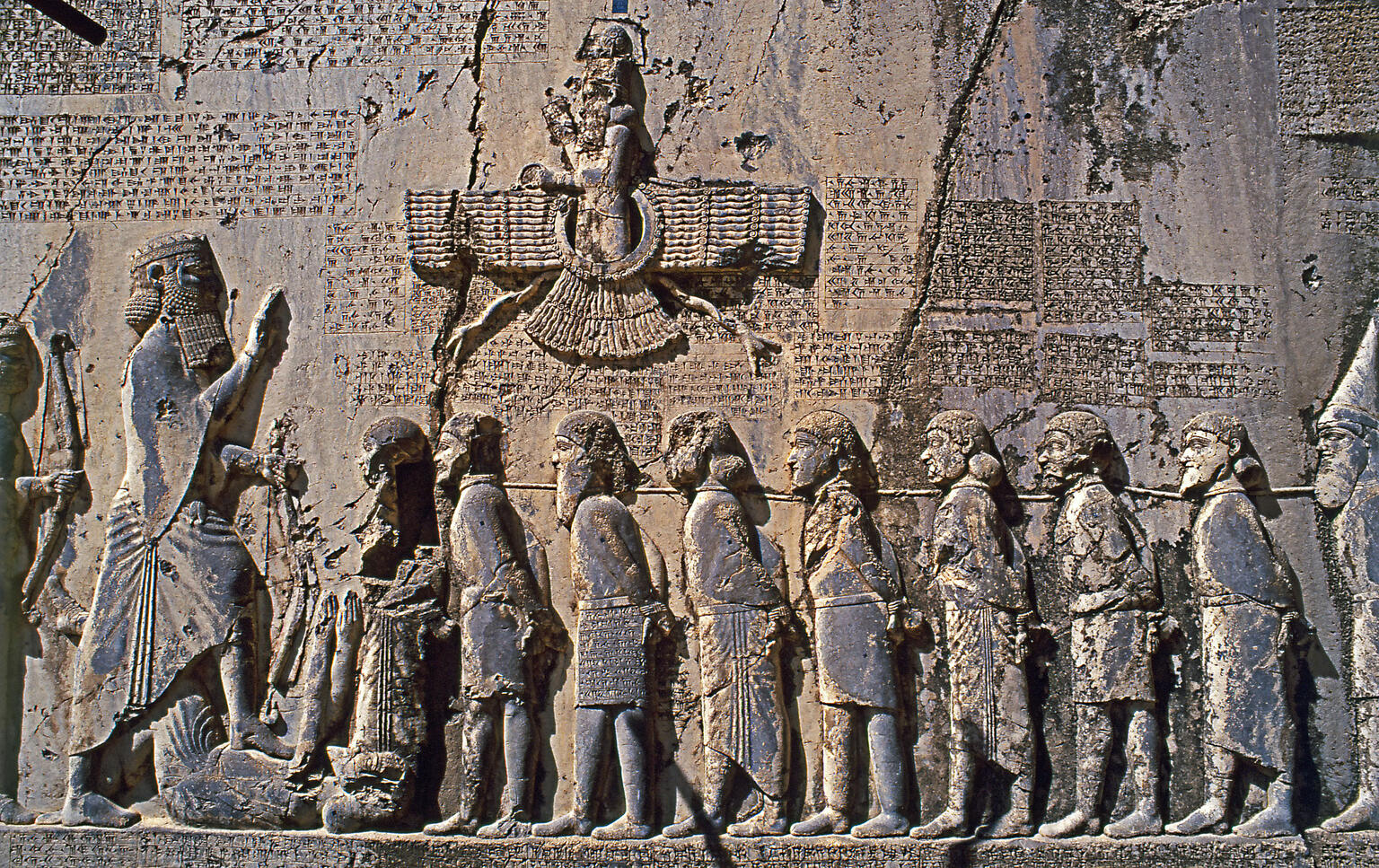 Relief with figures with hands bound standing next to larger figure along wall with relief of winged figure and Persian, Elamite, and Akkadian inscriptions.