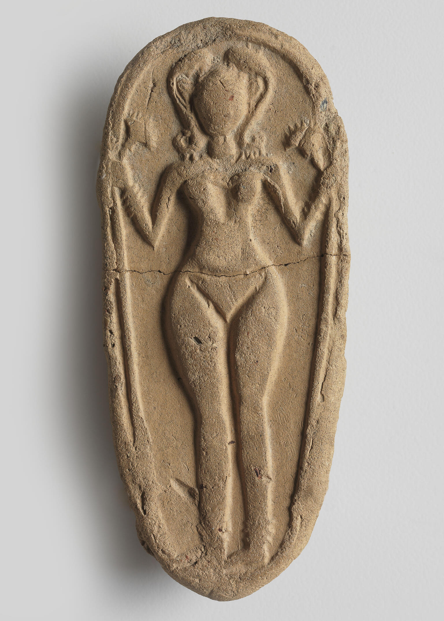 Oval clay piece with figurine of naked woman decorated with headdress, necklace, bracelets, and anklets, and holding a lotus flower.